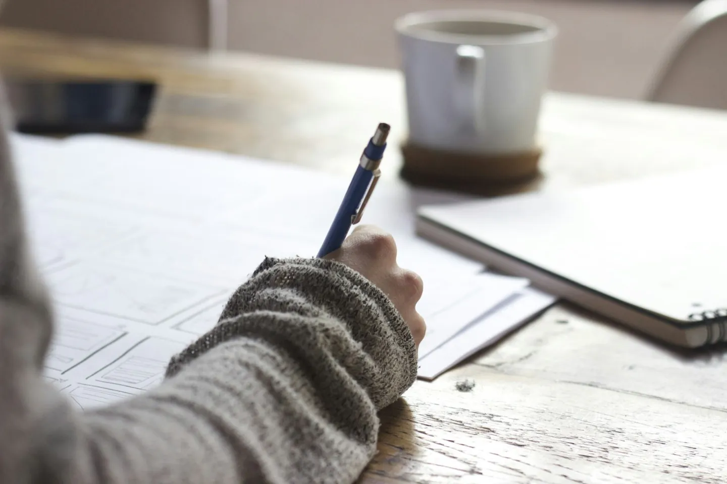A desk in soft daylight: notes half-written, a pen still in hand, a notebook and a cup set to one side.