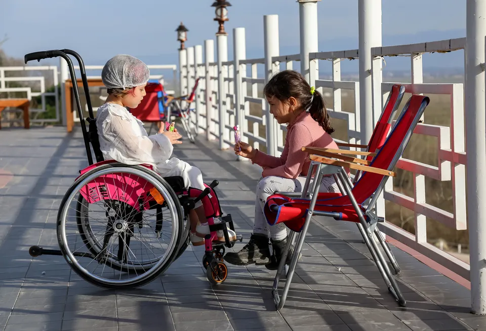 Two children playing with dolls outdoors, one seated in a wheelchair.