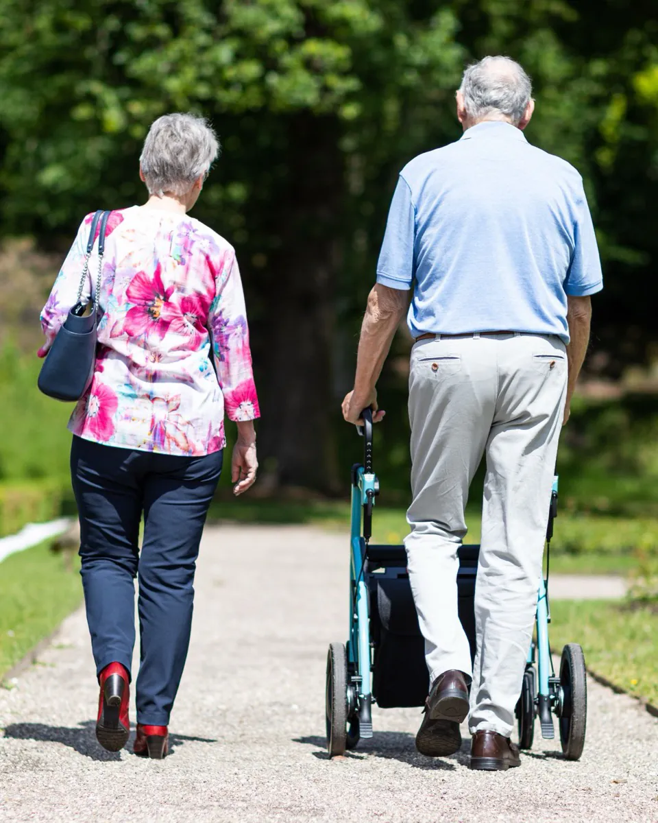 An older couple walking away down a park path, he with a wheeled walker.