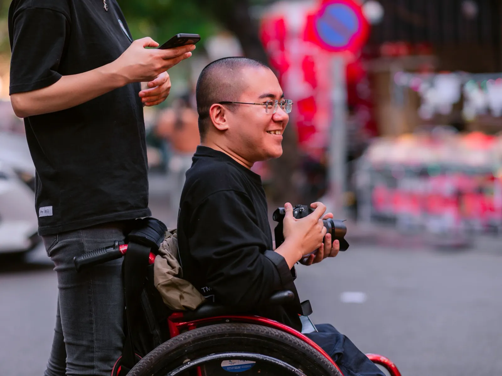 A smiling man in a wheelchair on a busy street, holding a camera.