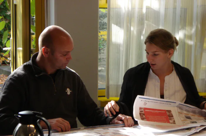 Two people side by side at a table by a window, going through a printed handout together while one of them explains.